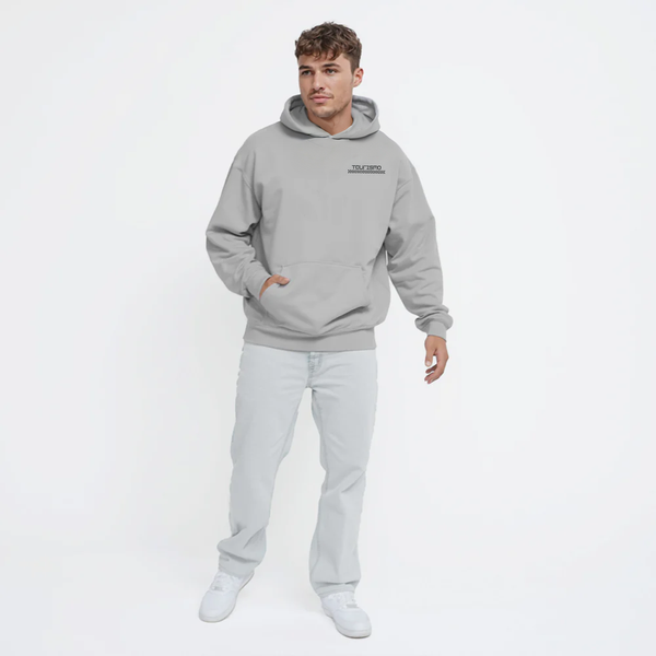 Homme portant un hoodie gris clair TOURISMO avec un jean clair et des sneakers, fond blanc