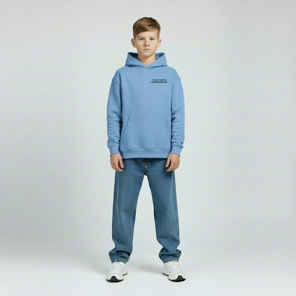Enfant portant le hoodie bleu Riviera Charles Leclerc, coupe ample, logo poitrine visible, fond studio gris, jambes légèrement écartées.