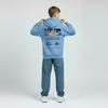 Enfant portant le hoodie bleu Riviera Charles Leclerc, coupe ample, logo poitrine visible, fond studio gris, jambes légèrement écartées.